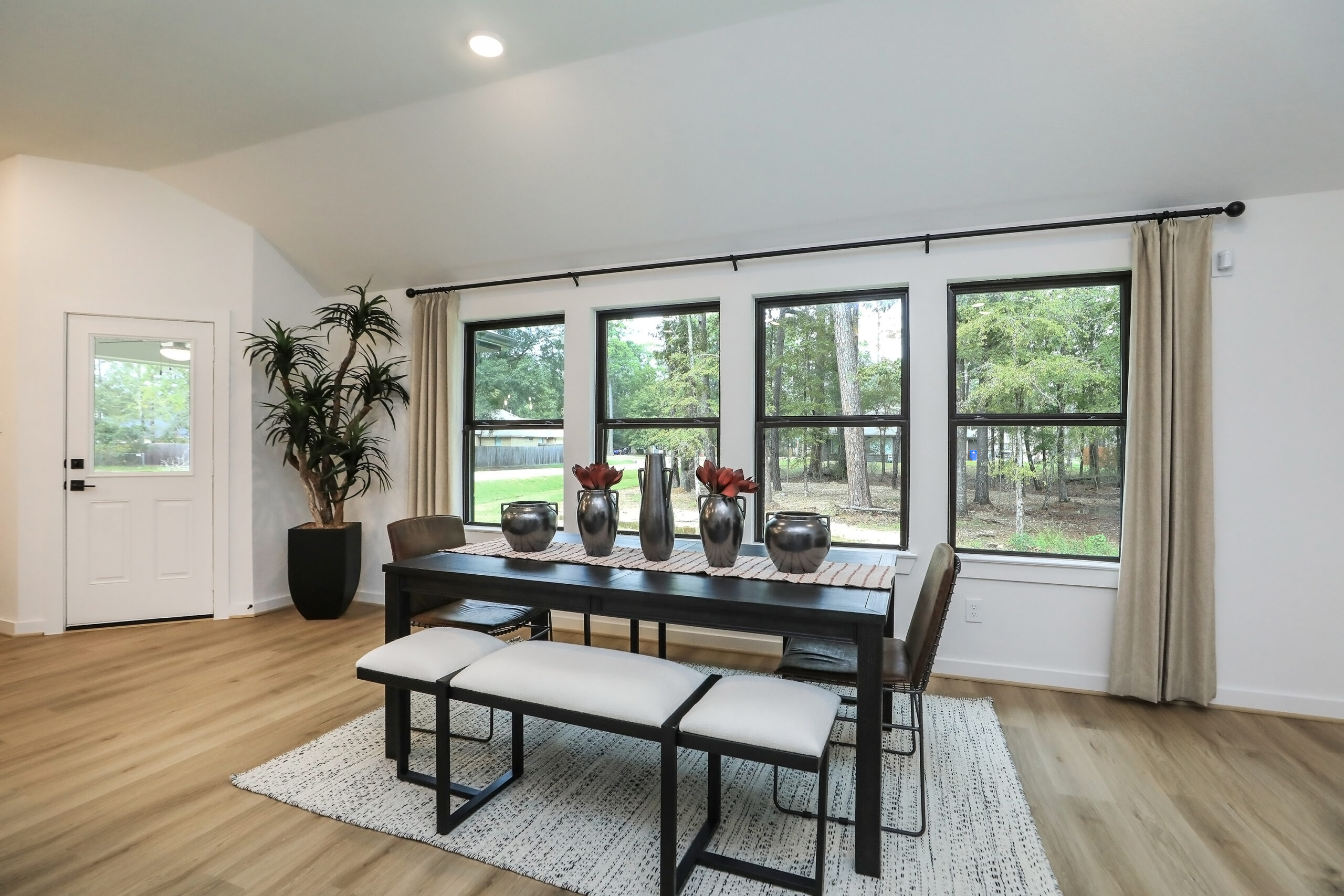 Modern dining room with large windows, a dark wood table, cushioned benches, decorative vases with red flowers, light hardwood floors, beige curtains, and a potted plant in the corner. Natural light fills the space.