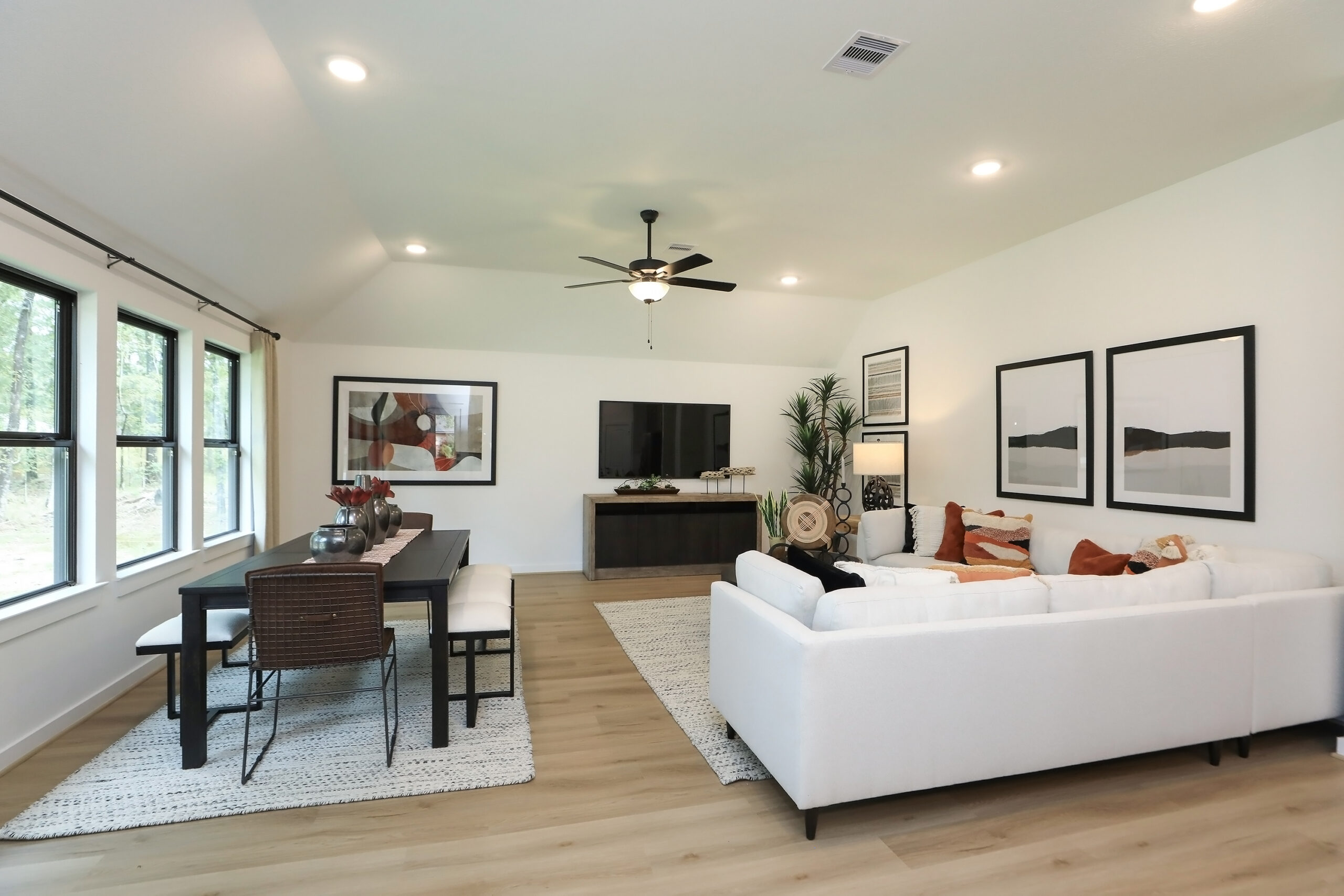 A modern living and dining area with light wood floors, a white sofa, a dining table with chairs, wall art, indoor plants, large windows, and a ceiling fan, all in a bright, open space.