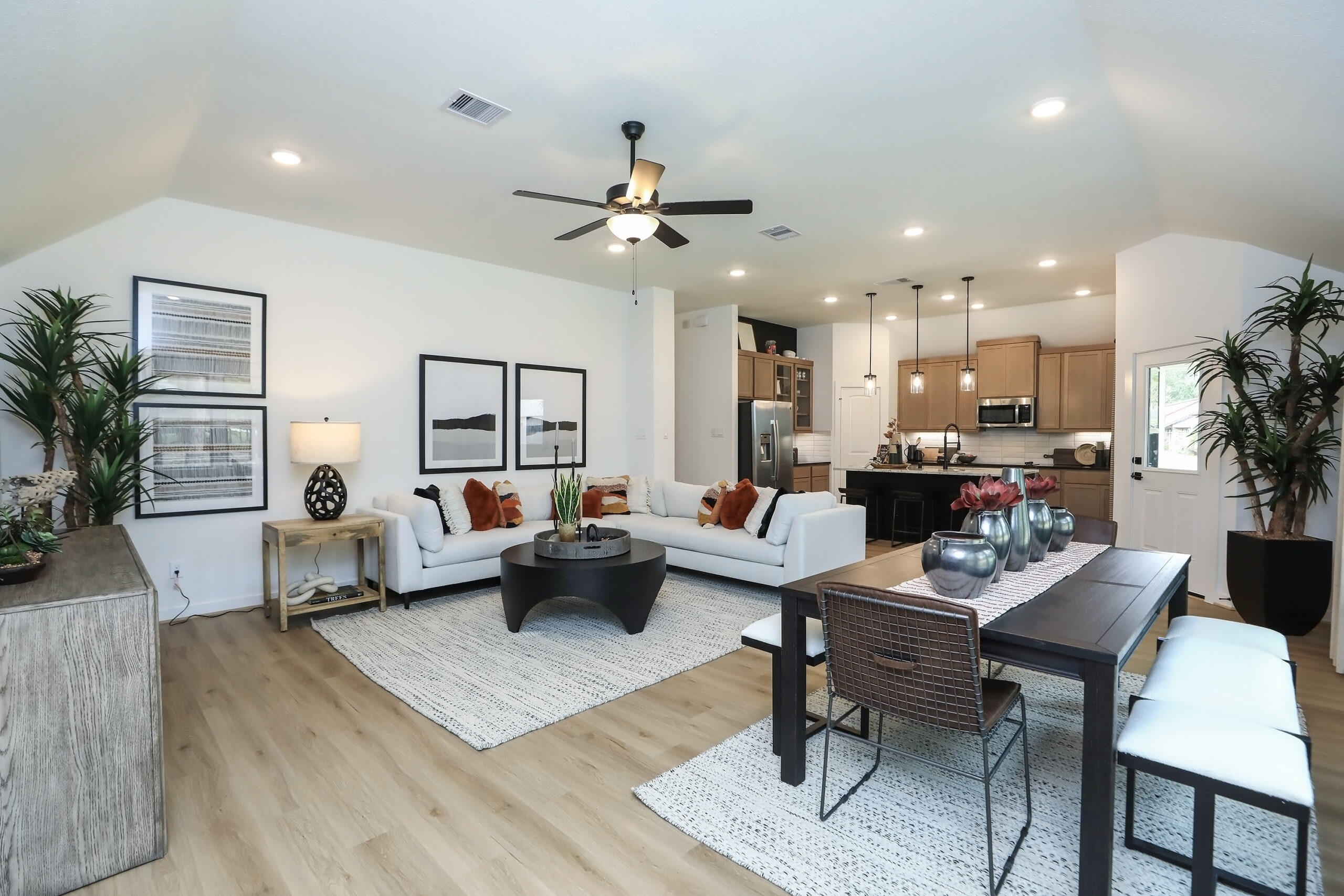 Modern open-concept living and dining area with light wood floors, white walls, neutral furniture, black accents, a ceiling fan, pendant lights, and kitchen in the background. Green plants and wall art add decorative touches.