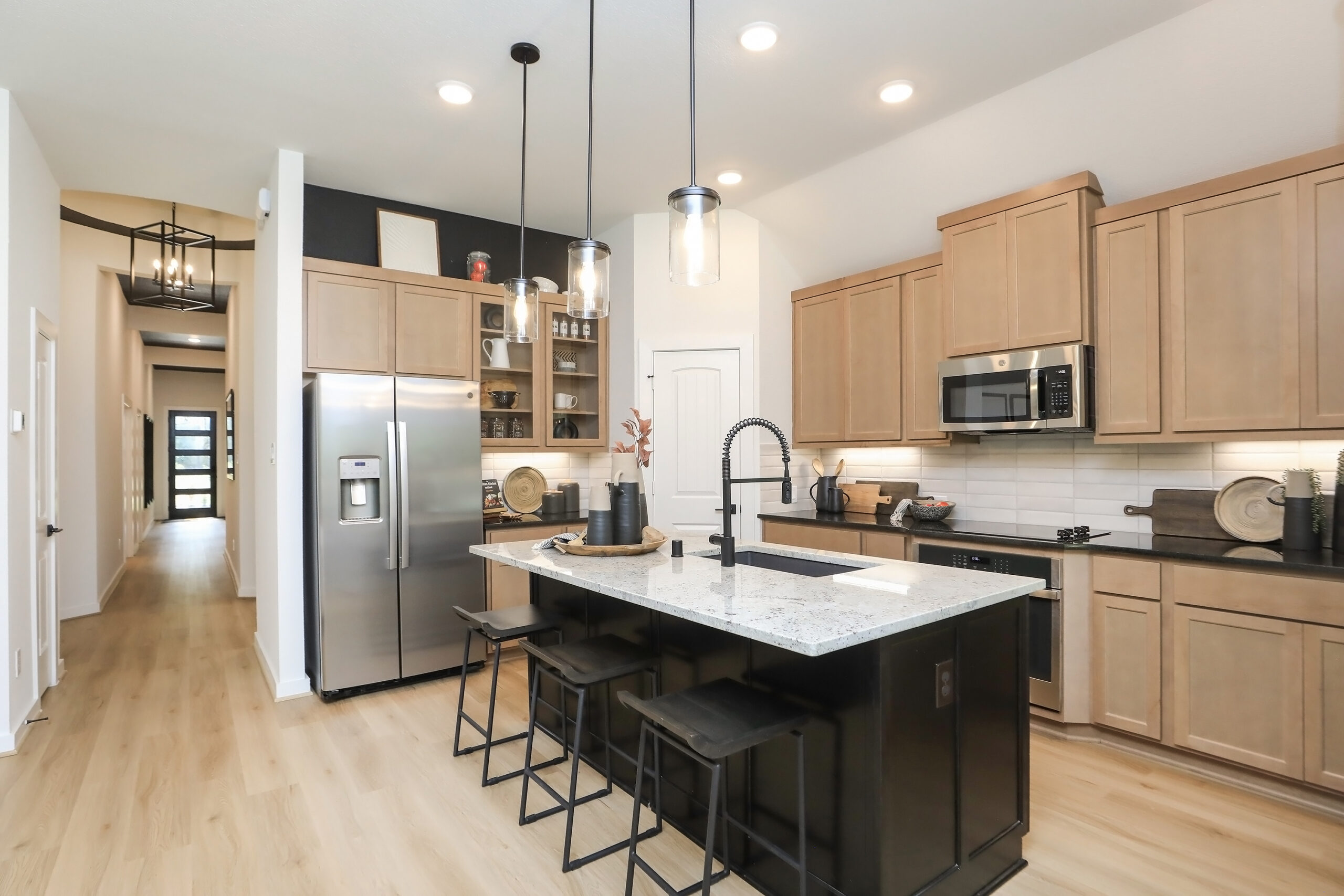 Modern kitchen with light wood cabinets, stainless steel appliances, a large island with a marble countertop, three black stools, pendant lights, and decorative items. Light wood flooring and a hallway are visible in the background.