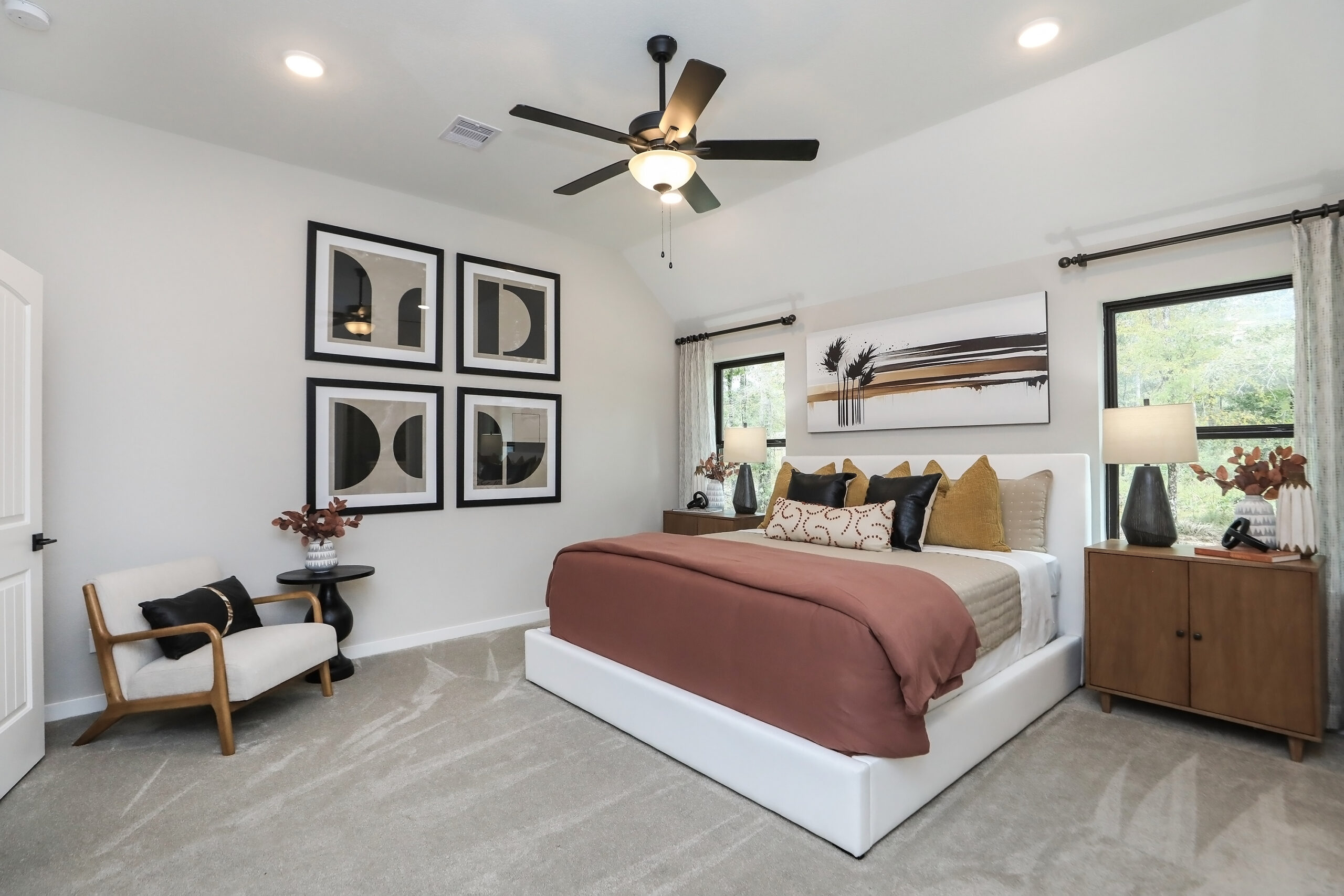Modern bedroom with a white bed frame, mauve comforter, decorative pillows, wooden nightstand with lamps, an armchair, large abstract wall art, ceiling fan, and two windows with curtains letting in natural light.