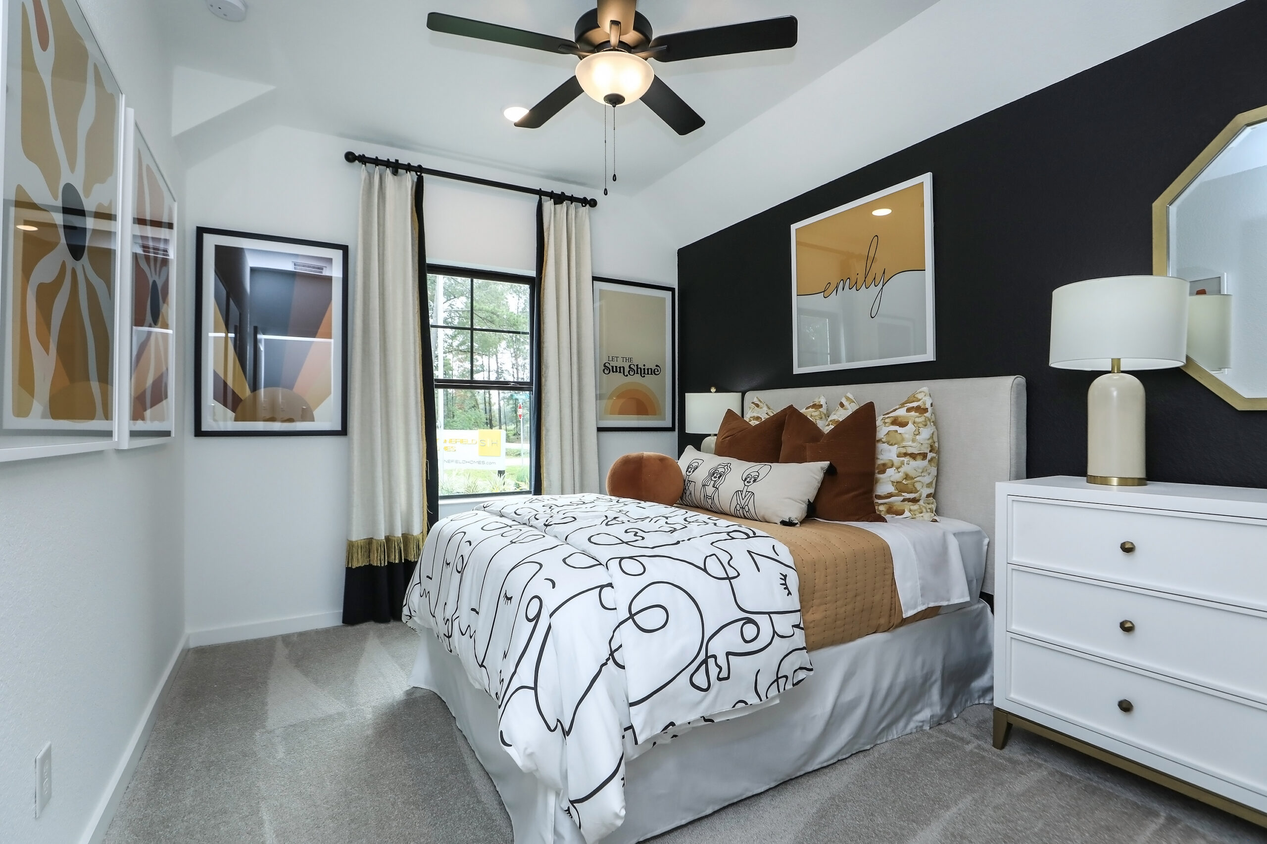 A modern bedroom with a bed featuring abstract black and white bedding, brown accent pillows, and contemporary wall art. A ceiling fan, large window with curtains, and a white nightstand with a lamp complete the decor.
