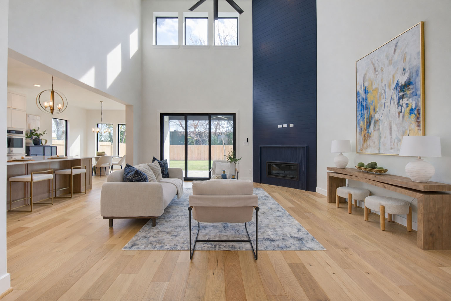 Modern living room with light wood floors, a beige sofa, a single chair, large blue accent wall with fireplace, abstract art, and high ceilings. Sunlight streams through tall windows and glass doors to a backyard.
