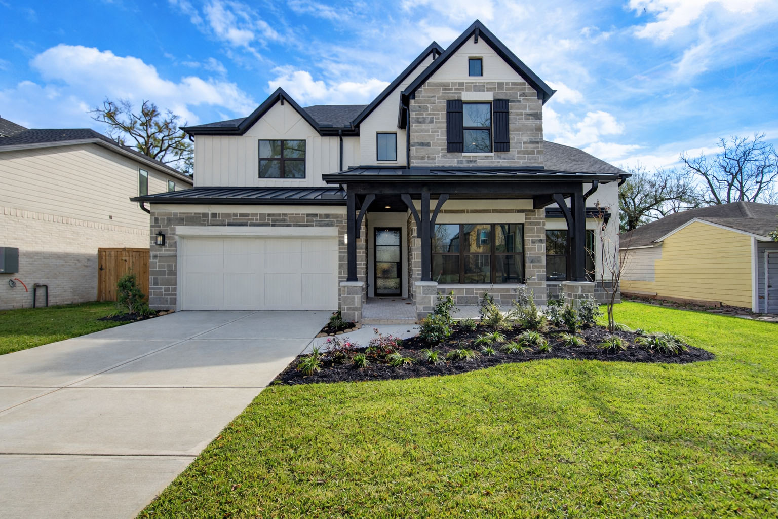 Two-story modern house with a stone and brick exterior, a covered front porch, attached garage, landscaped front yard, and a concrete driveway, set against a blue sky with scattered clouds.