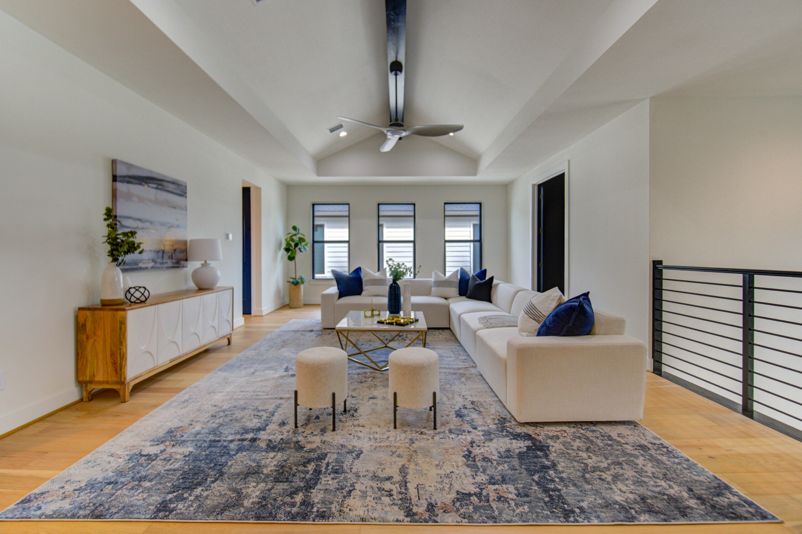 A modern living room with a vaulted ceiling, beige sectional sofa, blue and beige pillows, abstract rug, two round stools, a sideboard with decor, and large windows letting in natural light.