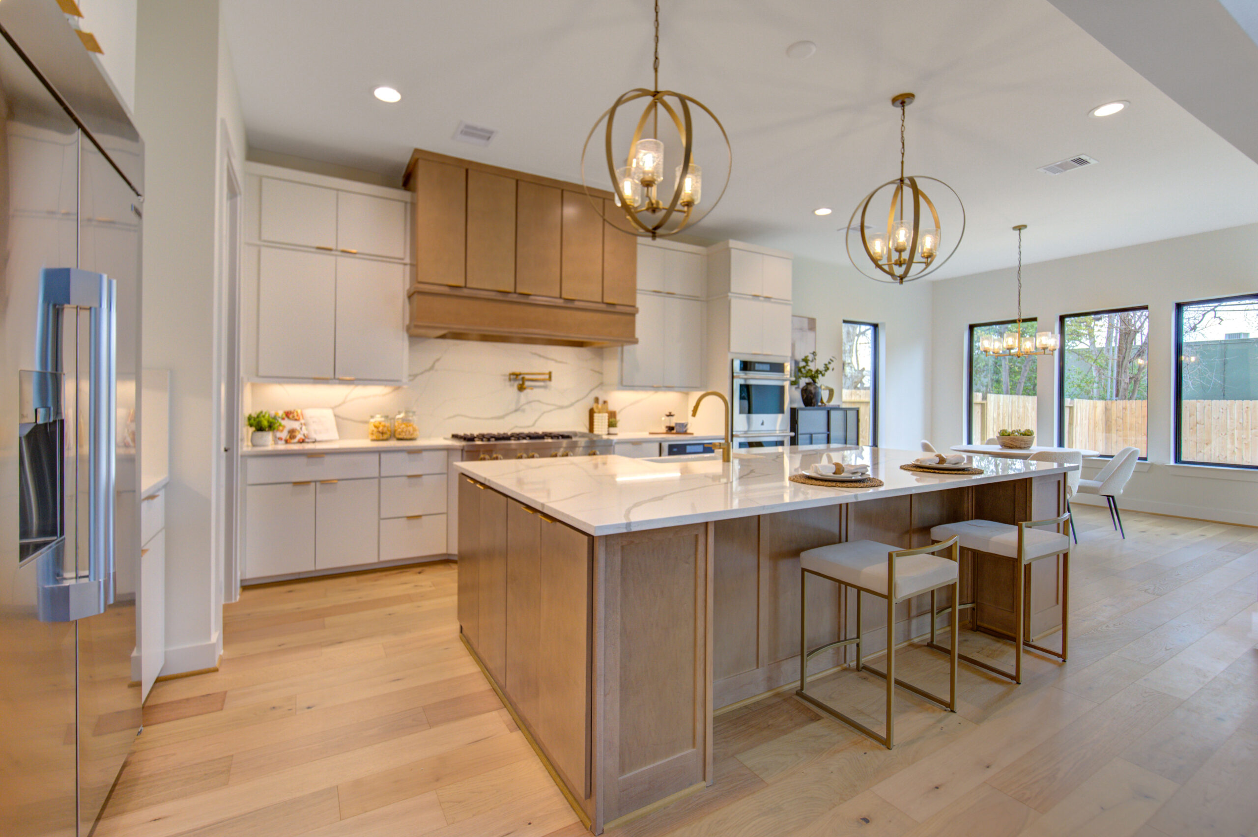 Modern kitchen with light wood floors, white cabinets, a large island with a marble countertop and wood base, gold pendant lights, barstools, built-in appliances, and large windows letting in natural light.