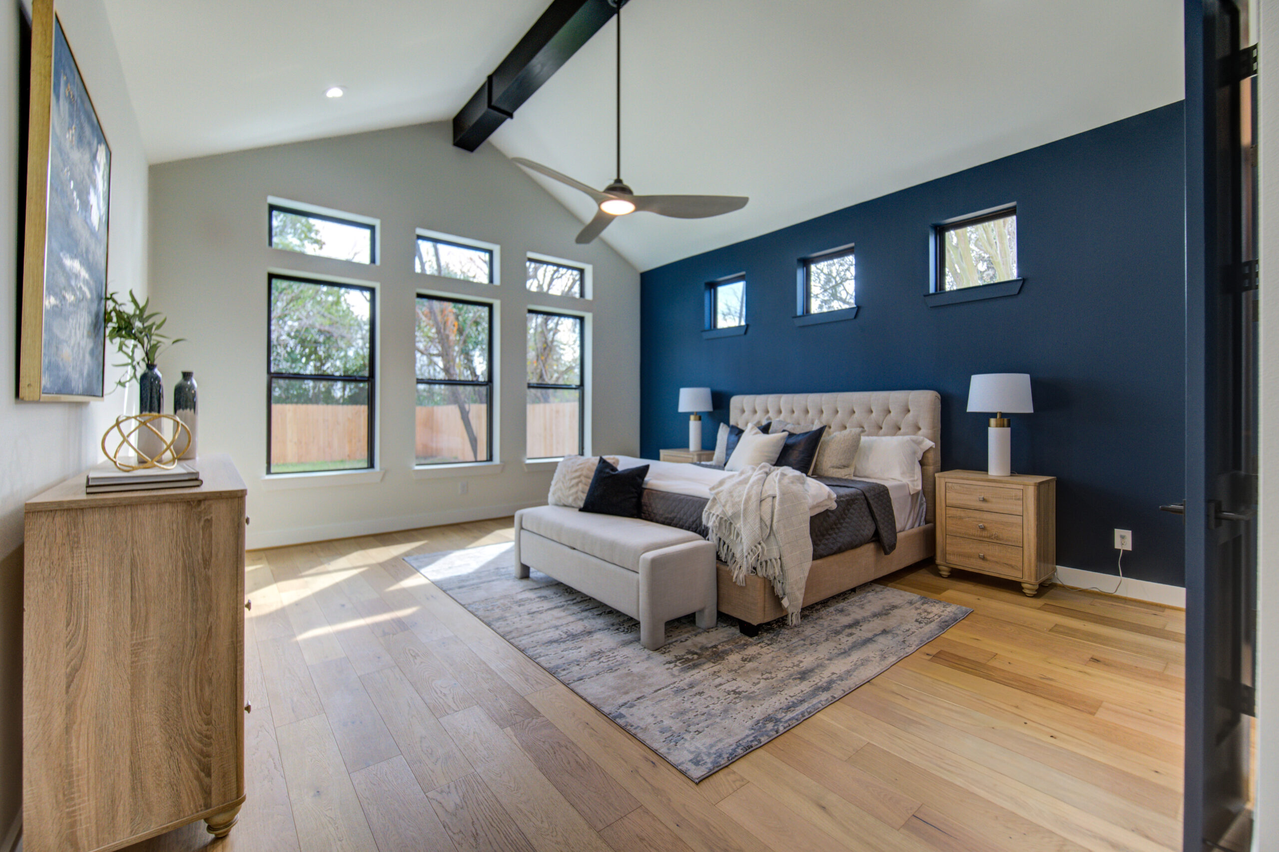 A modern bedroom with wood flooring, a dark blue accent wall, large windows, a tufted bed with gray and beige bedding, two nightstands with lamps, and a ceiling fan under exposed beams.