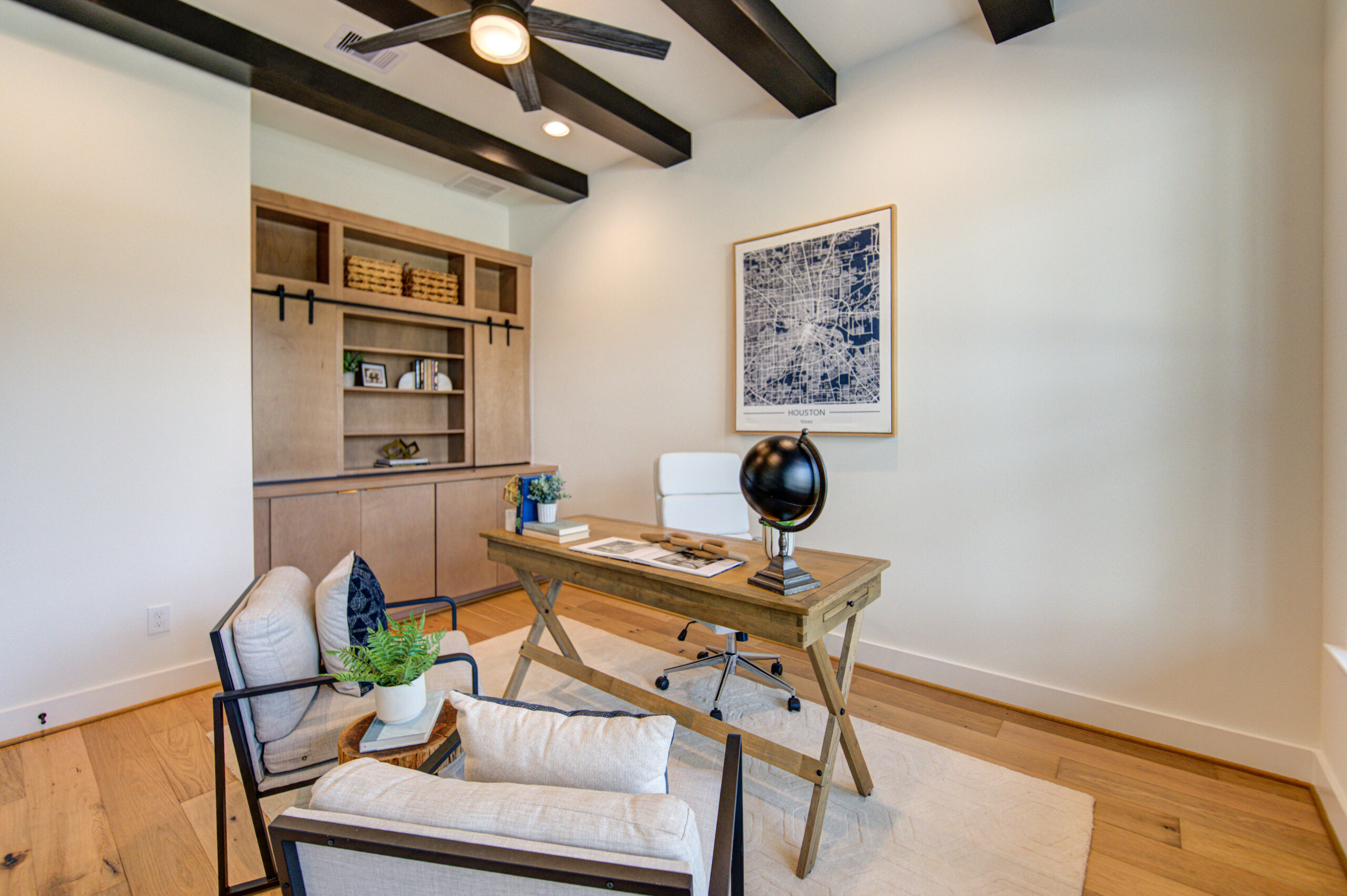 A modern home office with wood floors, a wooden desk with a globe and papers, a white office chair, a beige armchair, a potted plant, built-in shelves, and a large city map artwork on the wall.