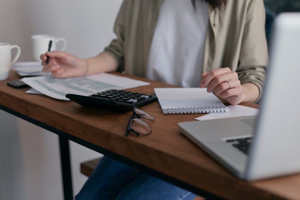A person sits at a desk with a laptop, calculator, notebook, documents, and glasses, appearing to work or study. Two coffee cups are also visible on the wooden table.