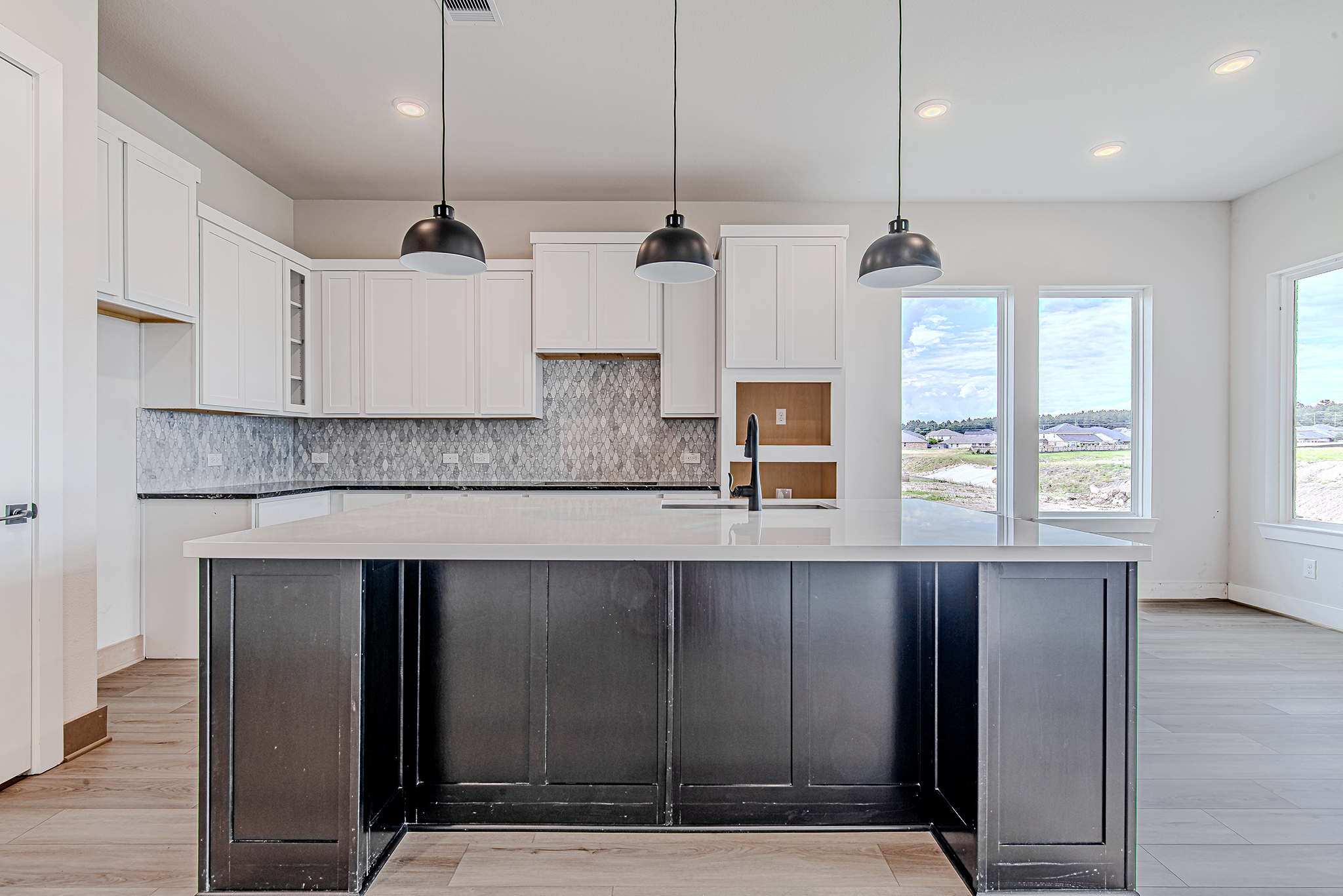 Modern kitchen with white cabinets, a large black island, three black pendant lights, gray tiled backsplash, wood-look flooring, and large windows letting in natural light with a view of a grassy landscape outside.