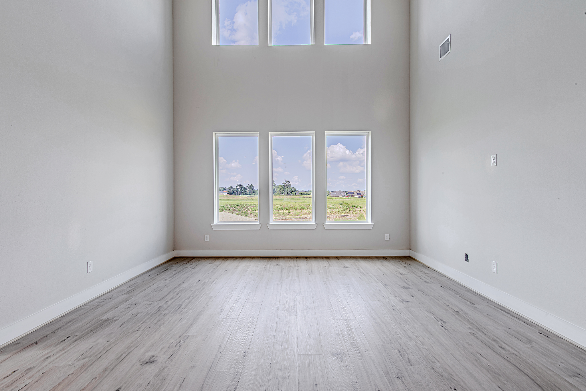 Spacious, empty room with light wood flooring, high ceiling, and large windows letting in natural light. The windows offer a view of a grassy landscape and a blue sky with clouds. Walls are painted white.