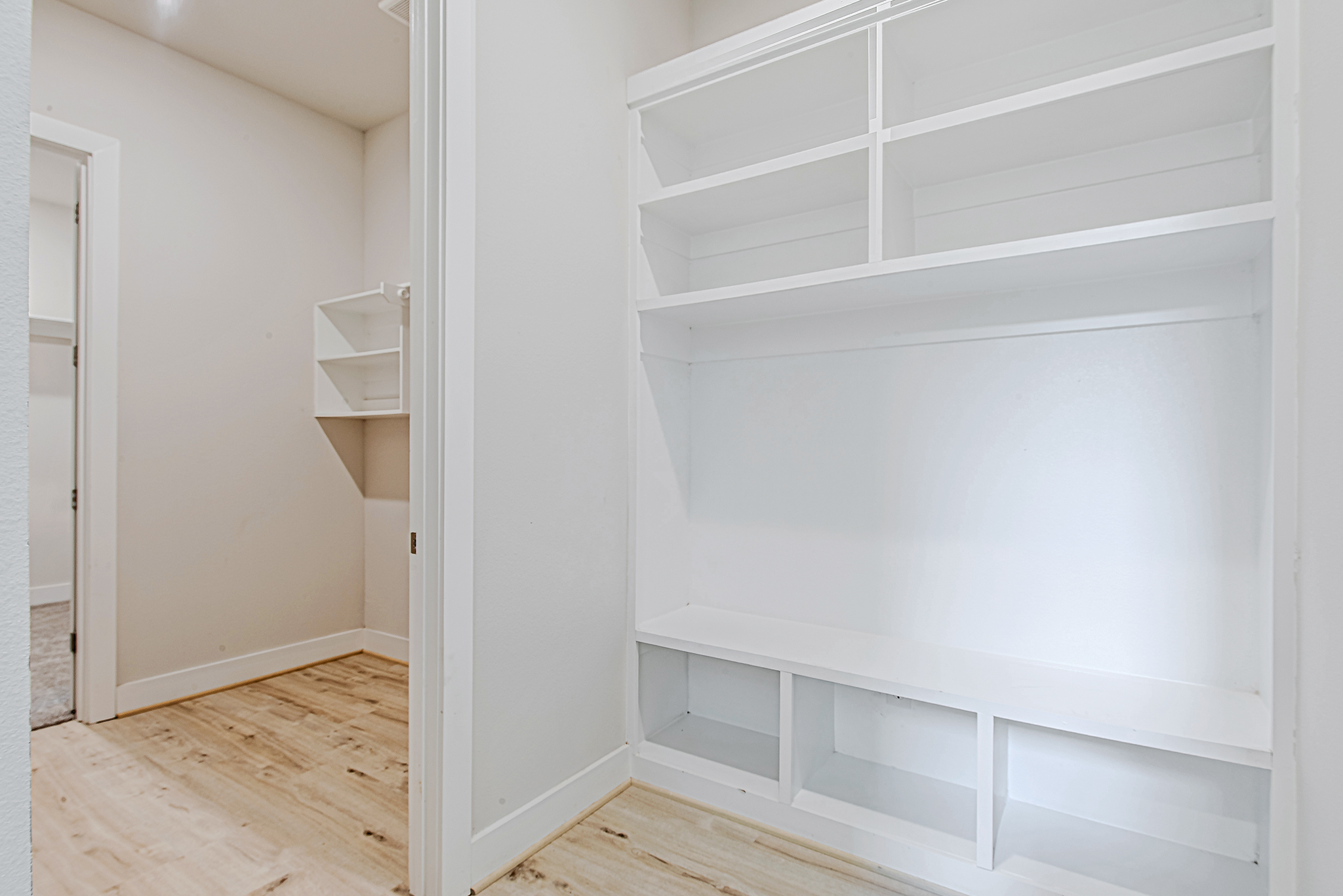 Walk-in closet with built-in white shelves and cubbies, light wood flooring, and neutral-colored walls; an additional shelving unit is visible through an open doorway.