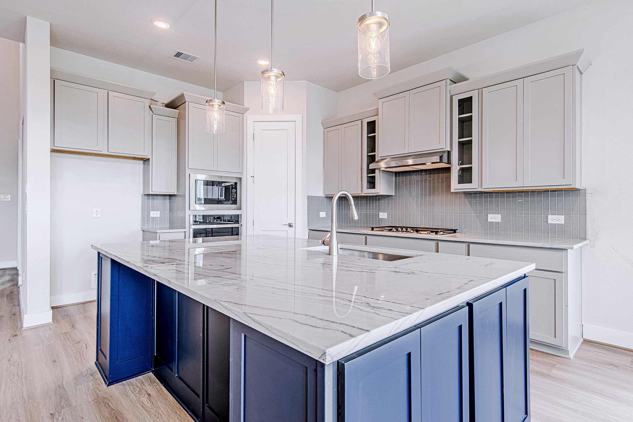 Modern kitchen with light gray cabinets, stainless steel appliances, a large marble island with a blue base, pendant lights above, light wood floors, and a neutral backsplash.