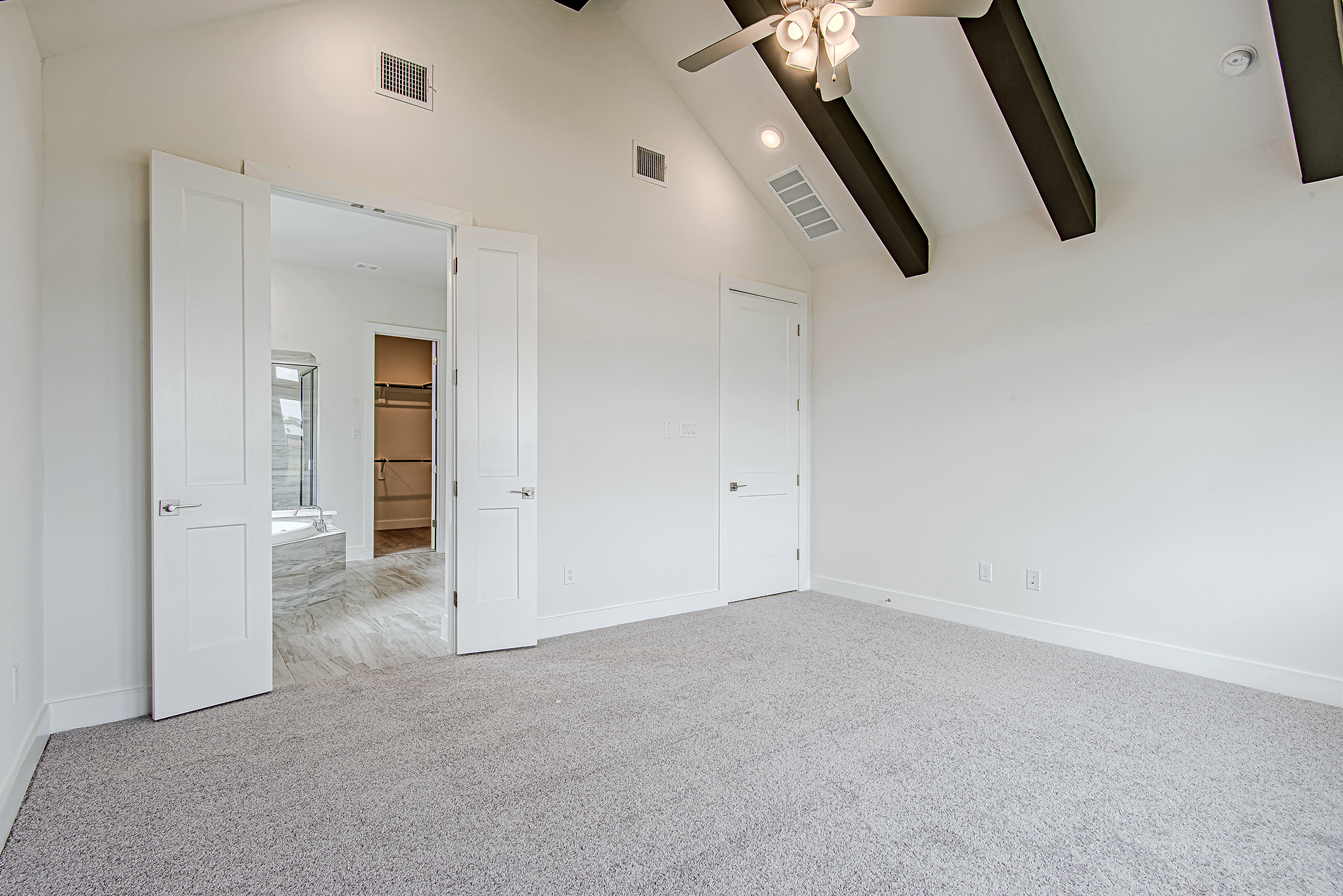 A bright, empty room with light gray carpet, white walls, black ceiling beams, a ceiling fan, and double doors leading to a bathroom with tile flooring.
