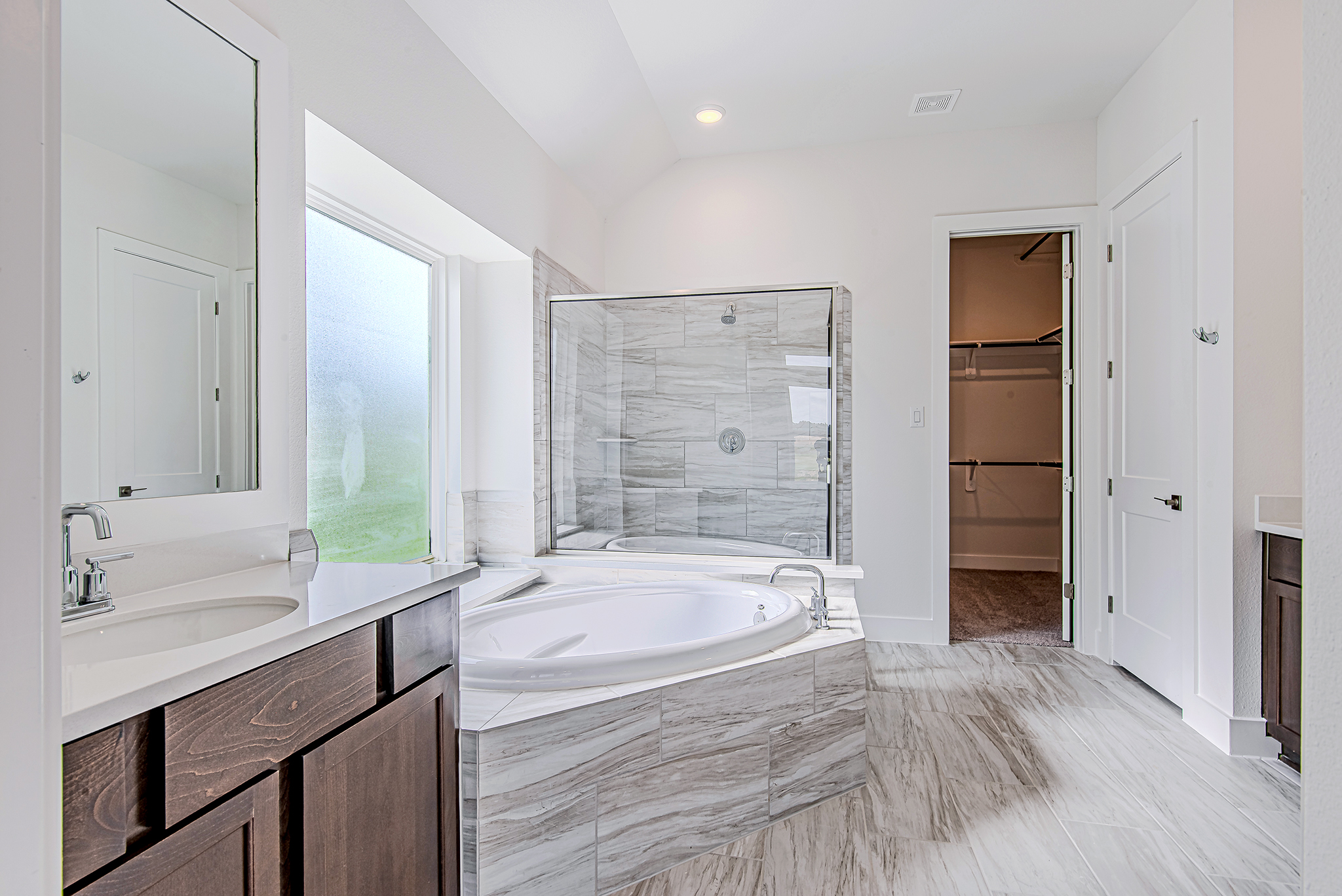 Modern bathroom with a large soaking tub, glass-enclosed shower, dark wood cabinets, white countertops, a mirror, and a walk-in closet with shelving visible through an open door. Natural light comes in through a frosted window.