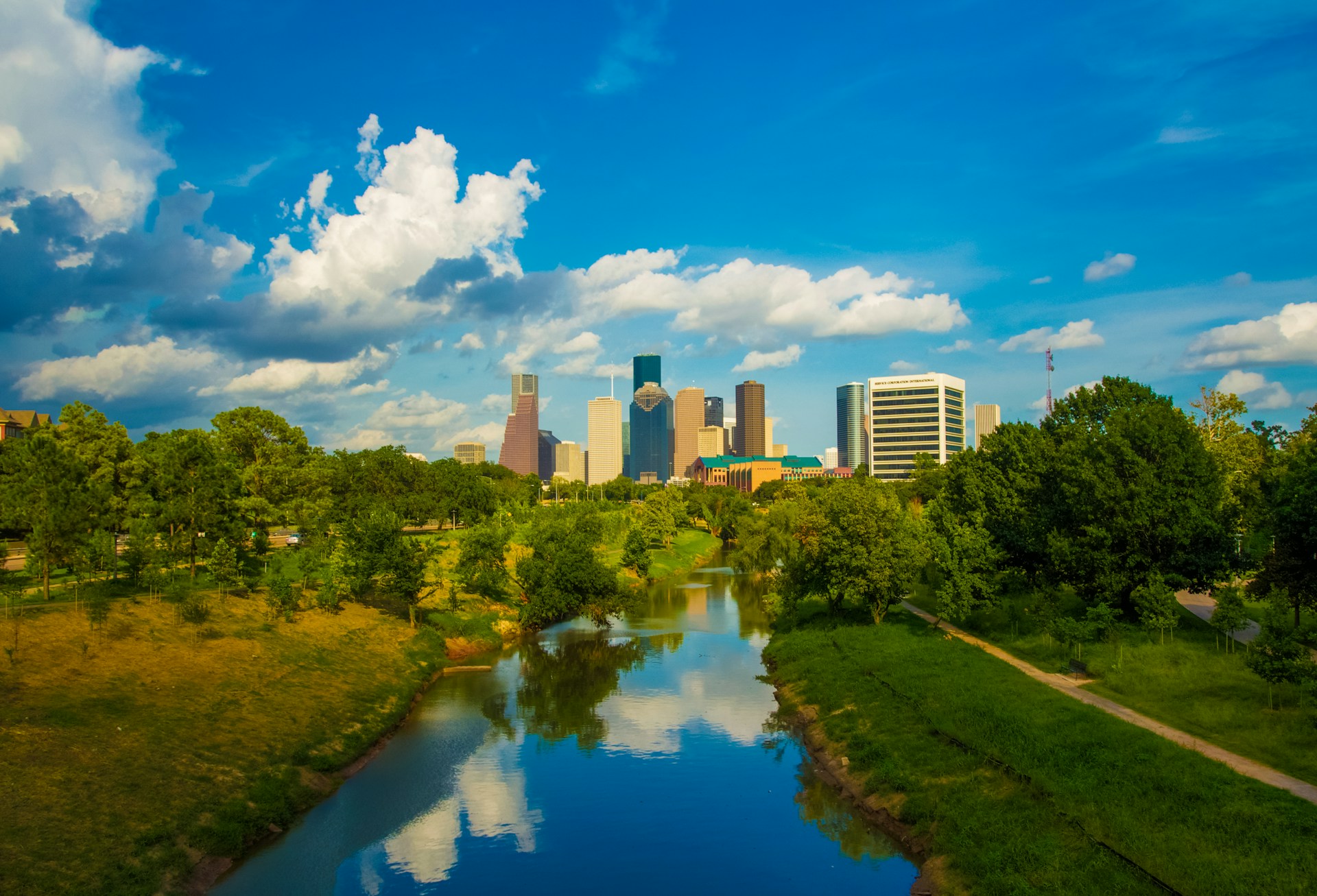 A river runs through a green park with trees on both sides, reflecting the blue sky and clouds. In the background, modern skyscrapers of a Houston skyline rise under a bright, sunny sky.