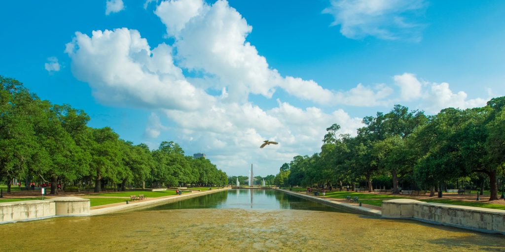 A reflecting pool stretches between tree-lined walkways under a bright blue sky with clouds. A bird flies above the water, and a tall monument stands in the distance.