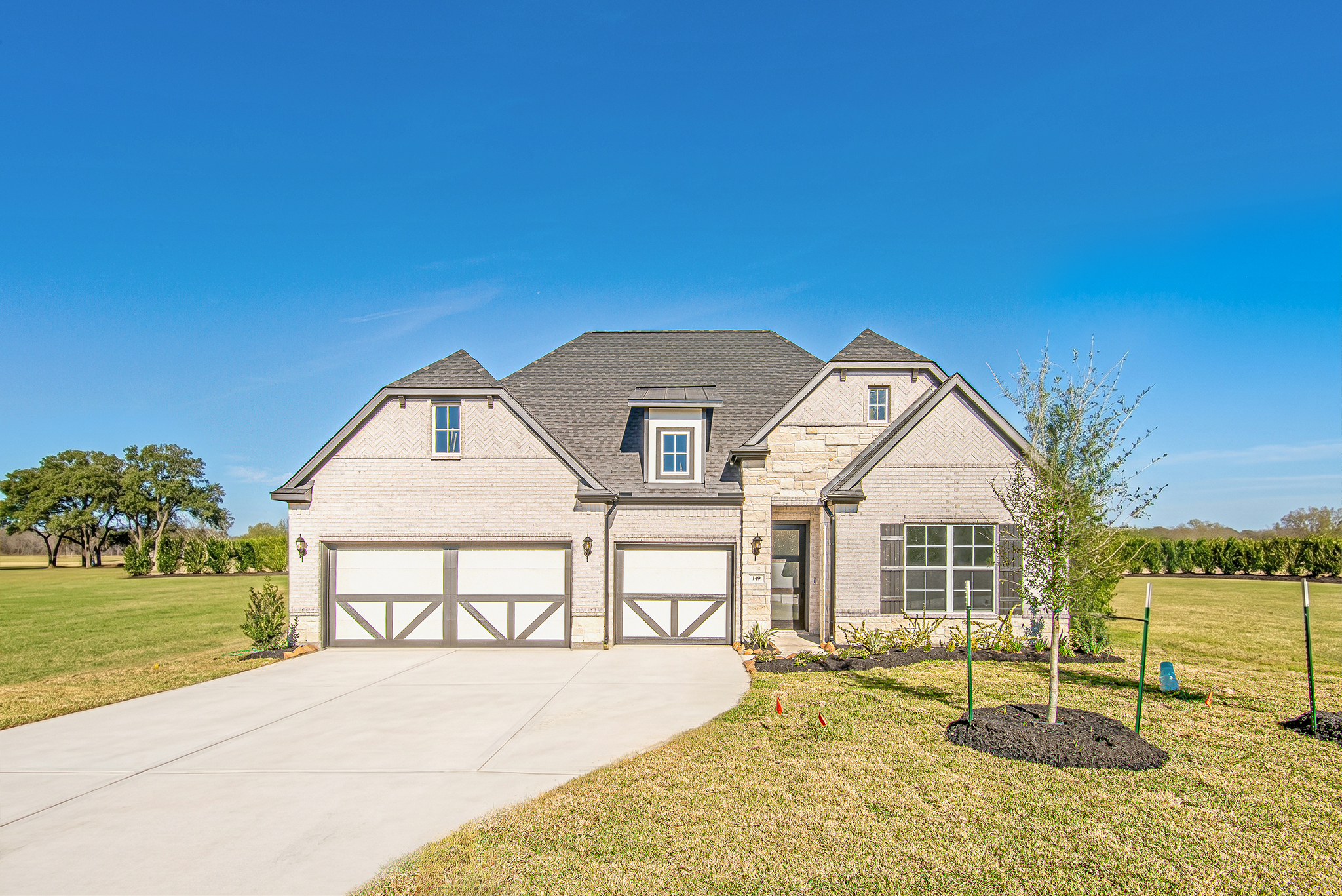 A modern, two-story brick house with three garage doors, a large driveway, and a neatly manicured lawn under a clear blue sky in a spacious, open area.