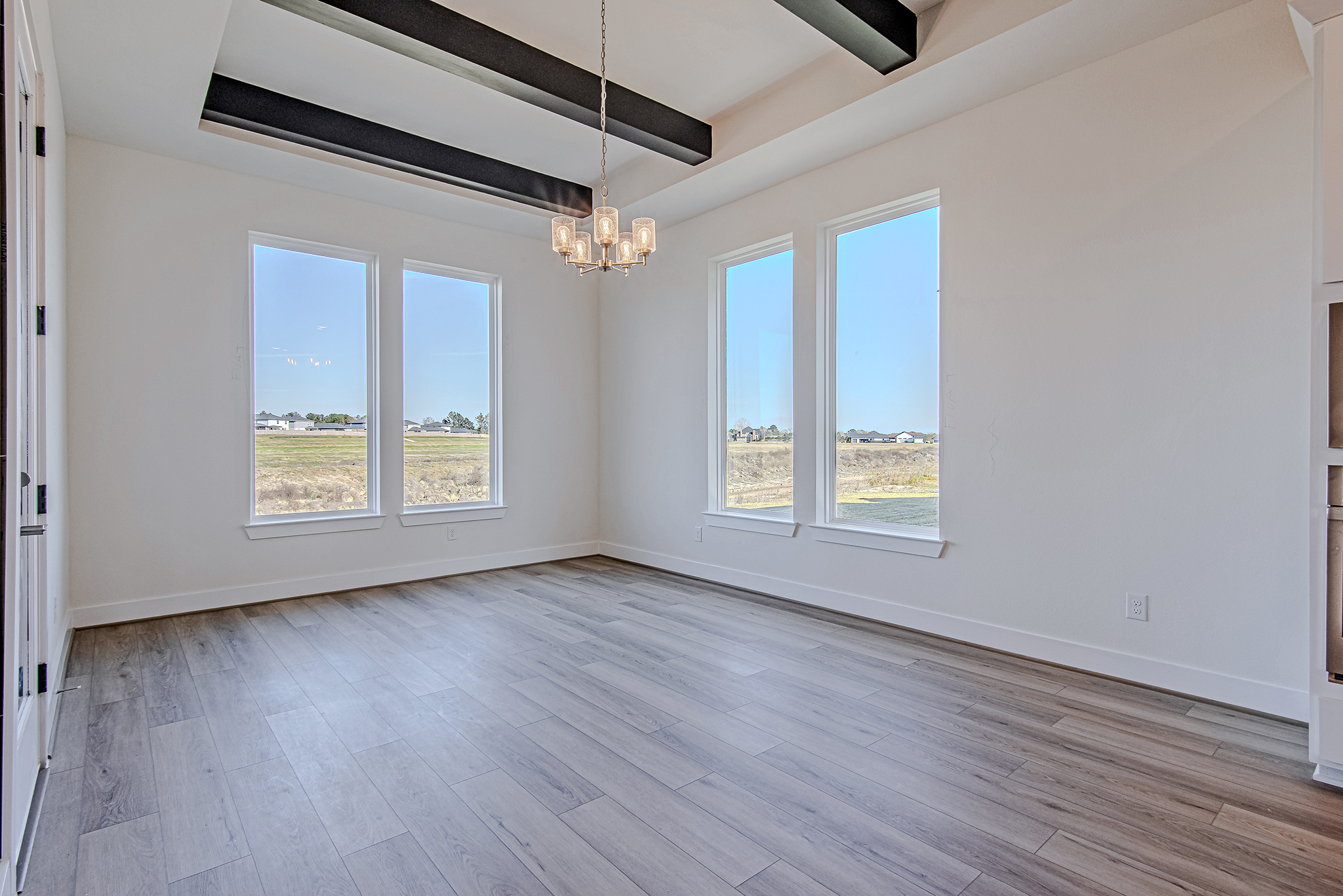 Bright, empty room with large windows, wood-like flooring, white walls, black ceiling beams, and a modern chandelier; outdoor landscape visible through the windows.
