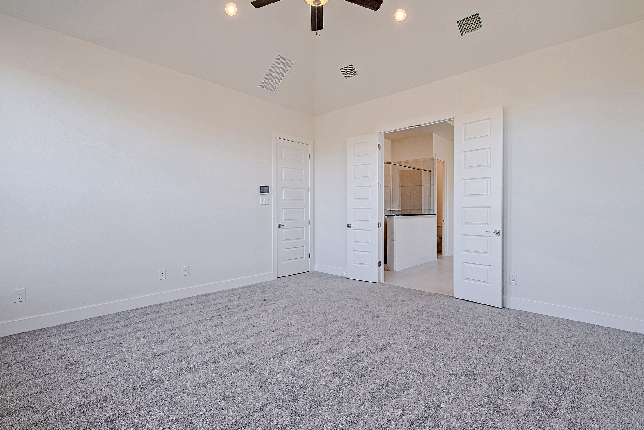 A bright, empty room with light-colored walls, gray carpet, a ceiling fan, and double doors leading to a bathroom with glass shower enclosure and tile flooring.