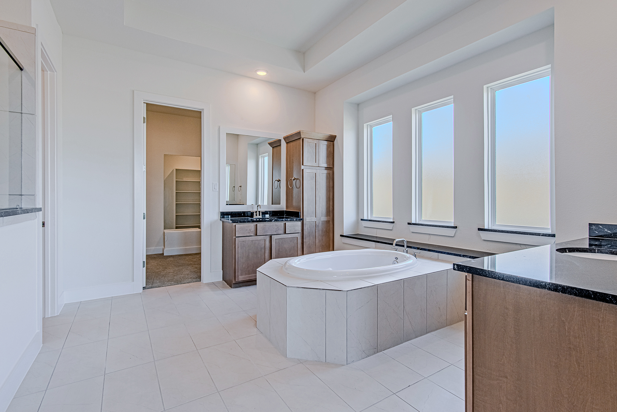 Spacious bathroom with a built-in oval bathtub, large windows, light tile flooring, dark countertops, wood cabinets, and an open doorway leading to a walk-in closet with shelving.