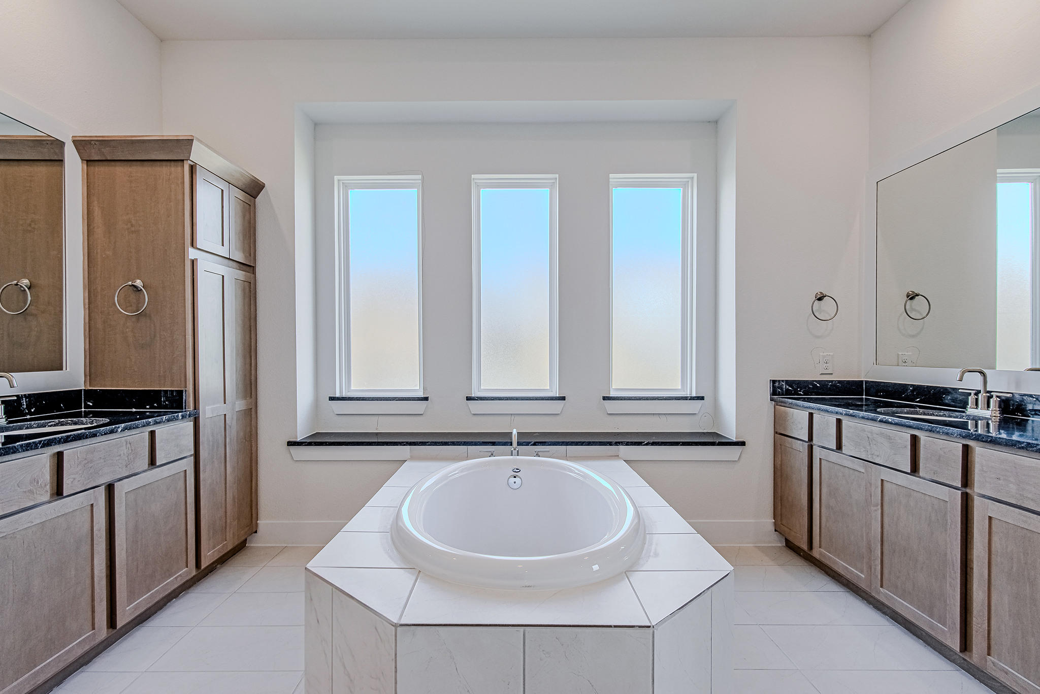 Modern bathroom with a central white bathtub, three frosted windows above, wooden cabinets and black countertops on both sides, and two mirrors on either wall. The space is bright and clean.