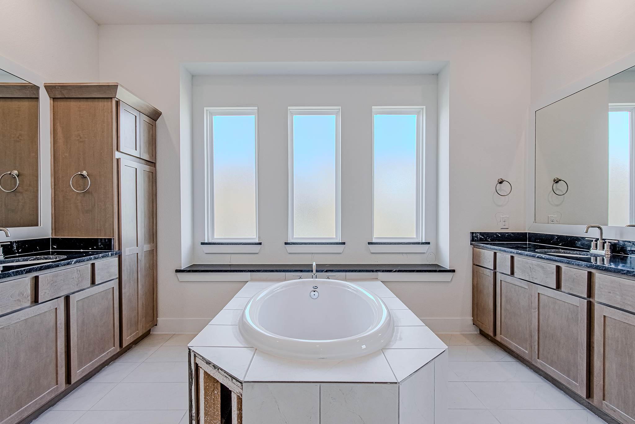 A modern bathroom with a central white bathtub, three frosted windows above, wooden cabinets on both sides, and large mirrors above black granite countertops.