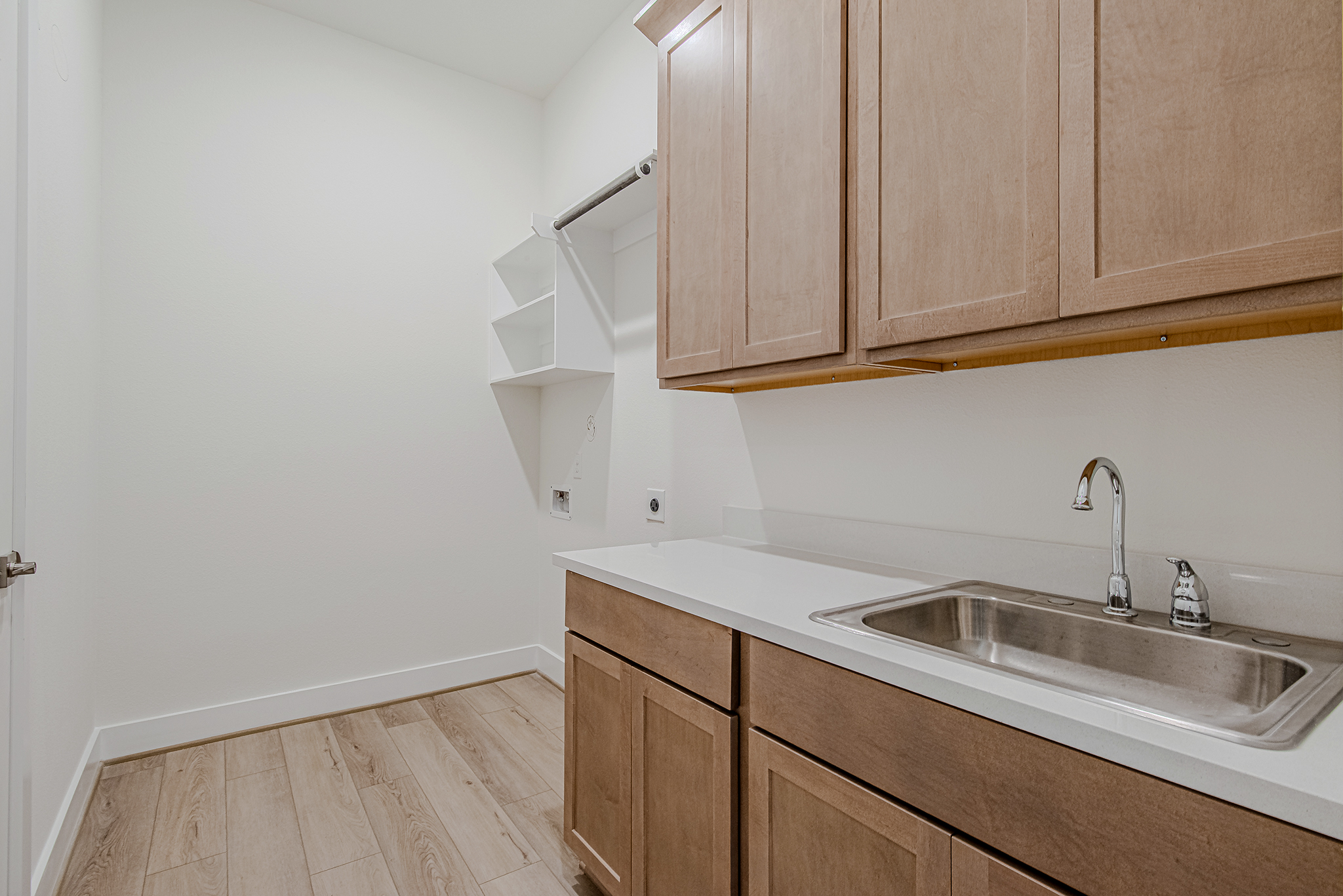 Laundry room with light wood cabinets, white countertop, a stainless steel sink with faucet, and open shelves on a white wall. The space has light wood flooring and a minimal, clean design.