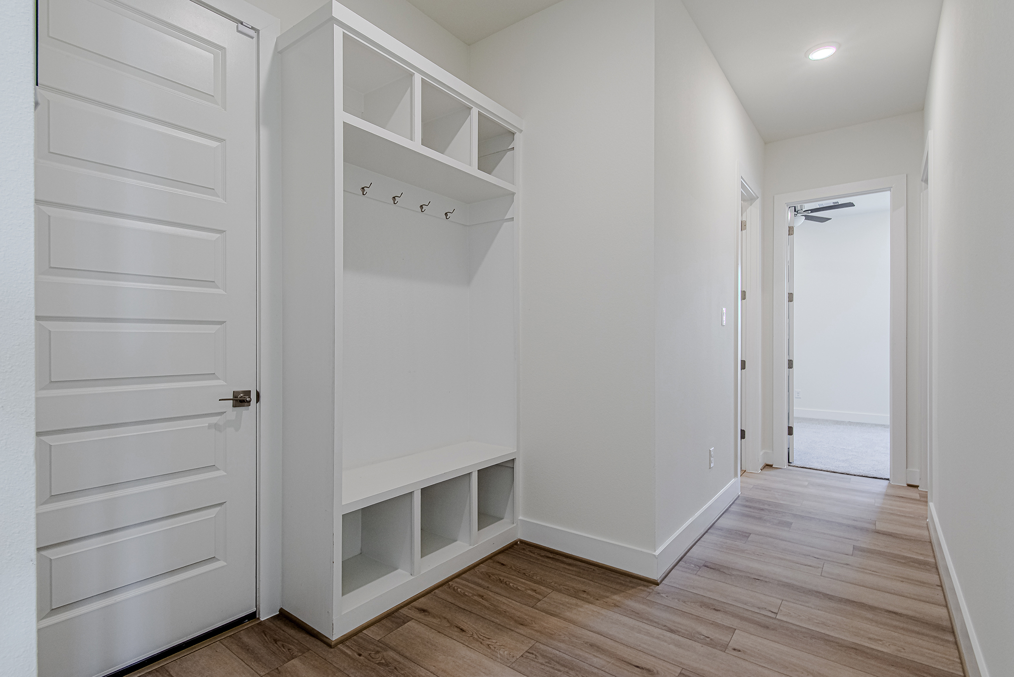 A modern hallway with light wood flooring, white walls, a white built-in storage bench with cubbies, coat hooks, and upper shelves. A white door is on the left, and a room with carpet is visible at the end.