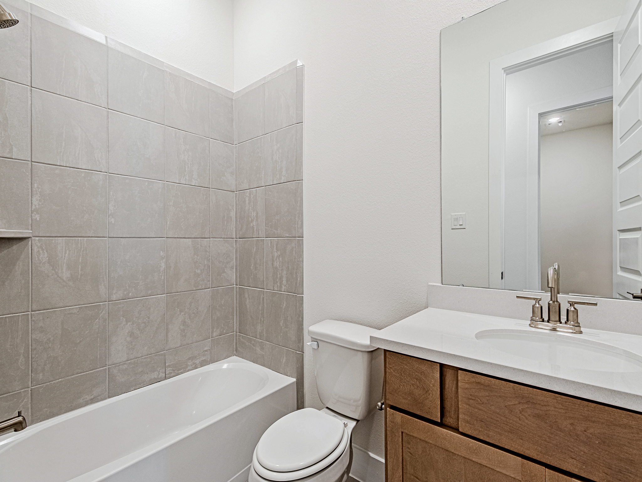 Modern bathroom with a tiled bathtub-shower combo, white toilet, wooden vanity with a white countertop, stainless steel faucet, and a large mirror above the sink. Walls are light-colored and the space looks clean and bright.