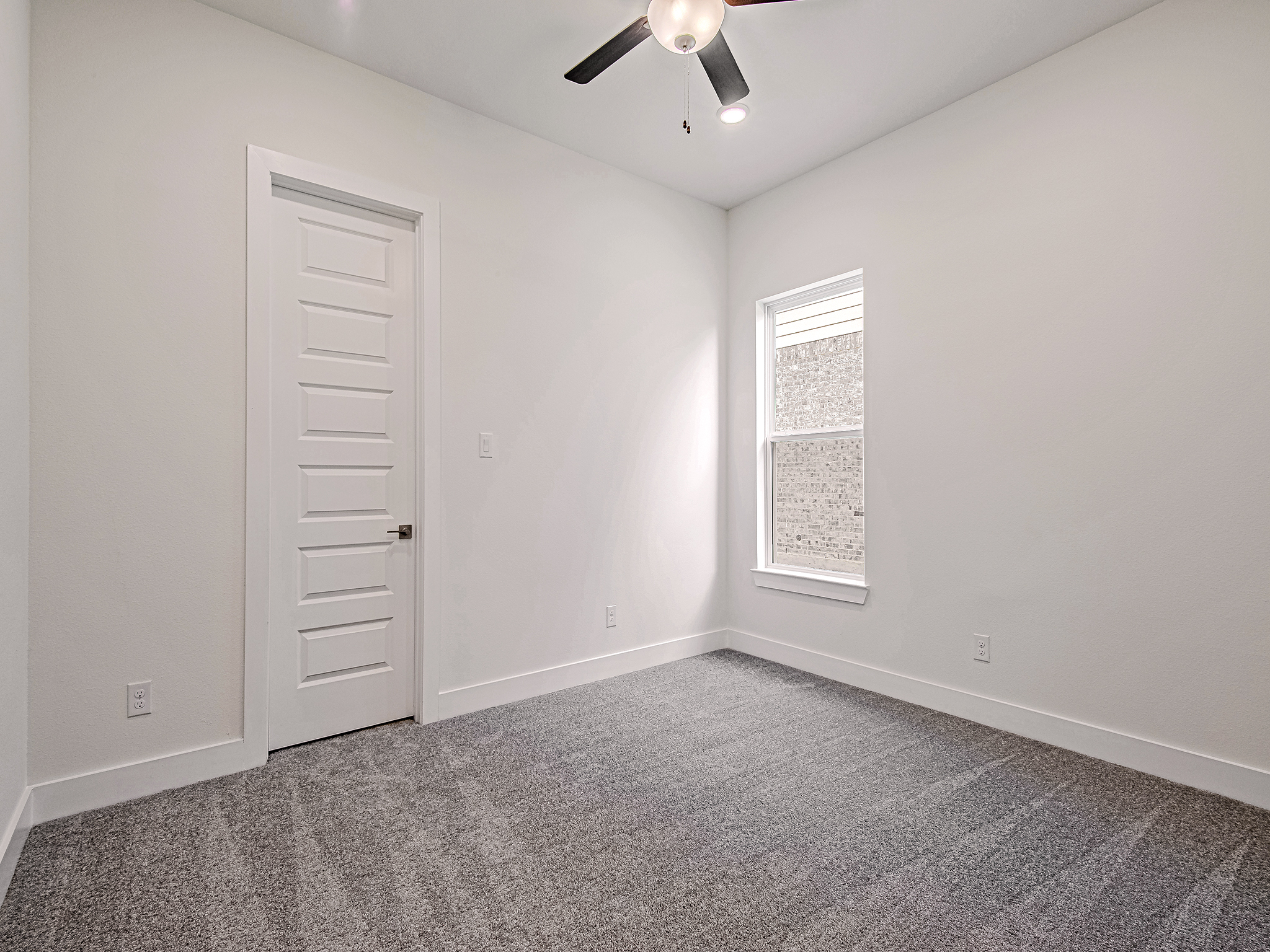 A minimal, empty room with light gray carpet, white walls, a white six-panel door, a large window letting in natural light, and a ceiling fan with three blades.