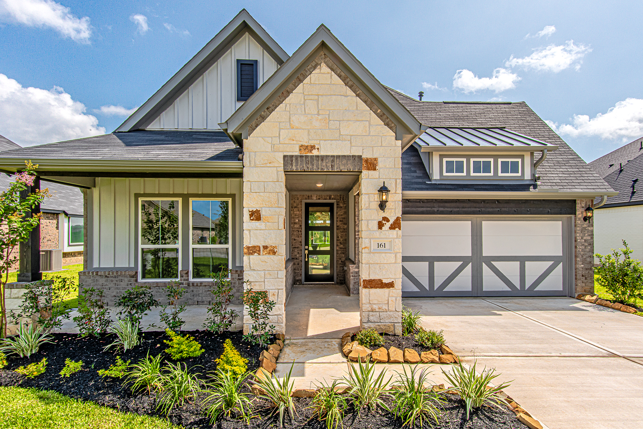 Modern suburban house with stone and siding exterior, covered front porch, double garage door, manicured lawn, and landscaped garden beds, set on a sunny day with blue skies and a few clouds.