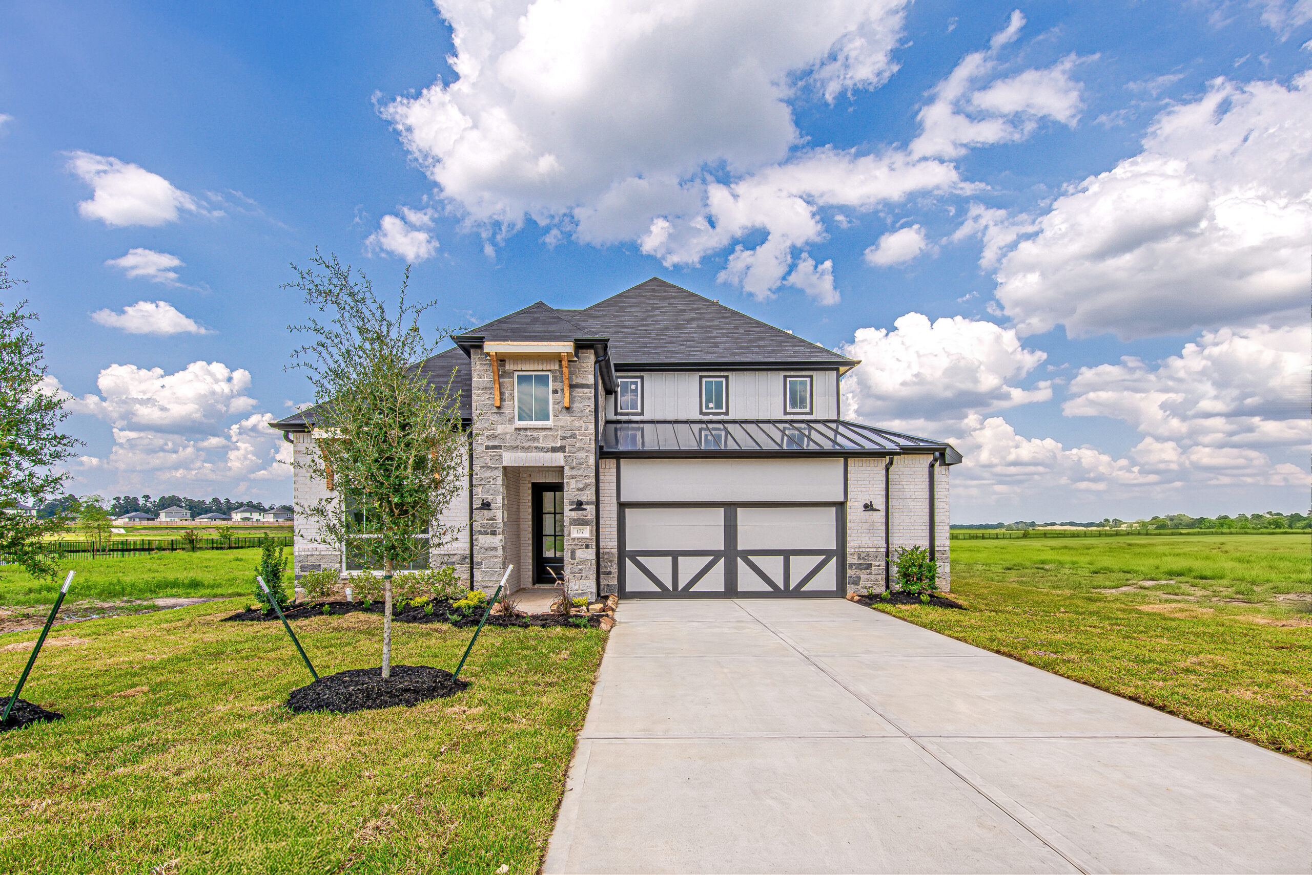 A modern two-story house with a gray roof, brick and stone exterior, double garage, and a long driveway, set on a spacious green lawn under a partly cloudy sky.