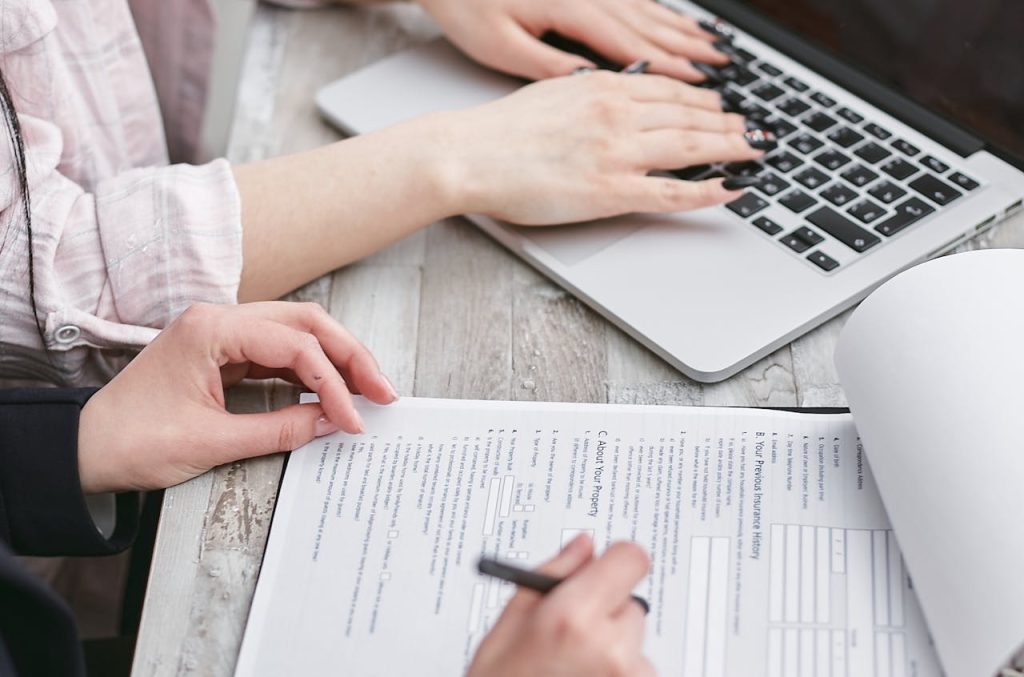 Two people work together at a desk; one types on a laptop while the other fills out a paper form with a pen. The workspace has a light wooden surface.