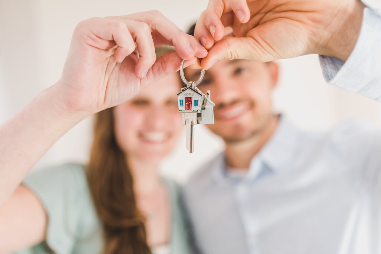 A smiling couple holds up a house-shaped keychain with a key, symbolizing buying a new home. The keychain is in focus, while the couple is blurred in the background.