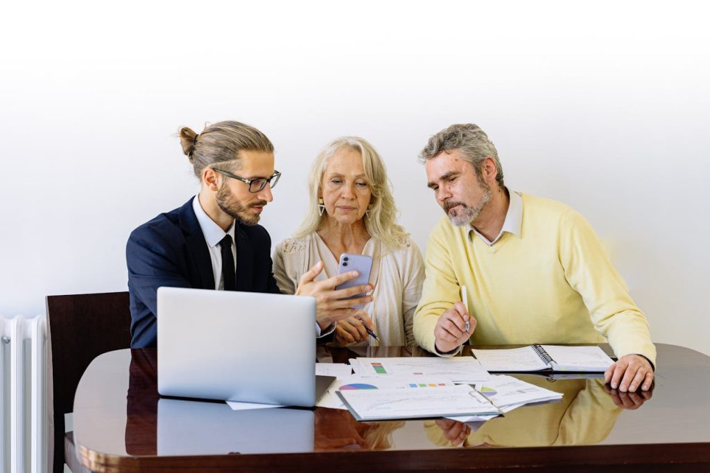 A young businessman shows a smartphone to an older couple as they sit together at a table with a laptop, documents, and charts, appearing to discuss financial or business matters.