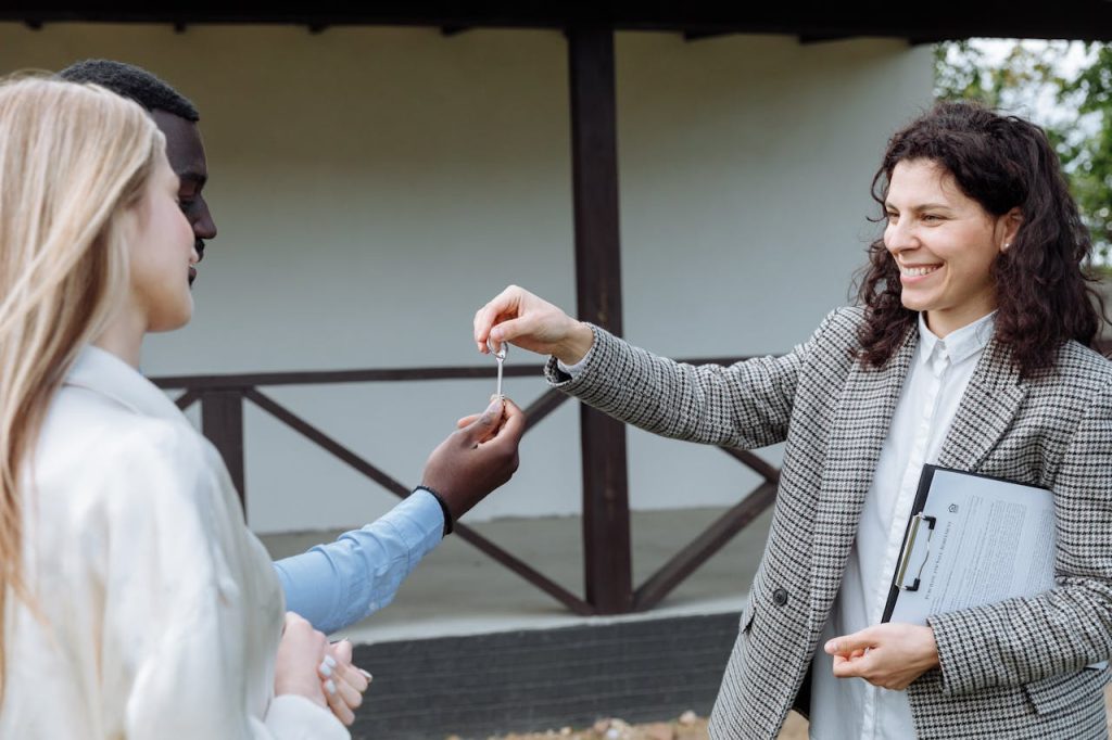 A smiling woman hands a key to a couple standing outside a house. The woman holds a clipboard and the couple appears happy, suggesting a real estate transaction or new home purchase.
