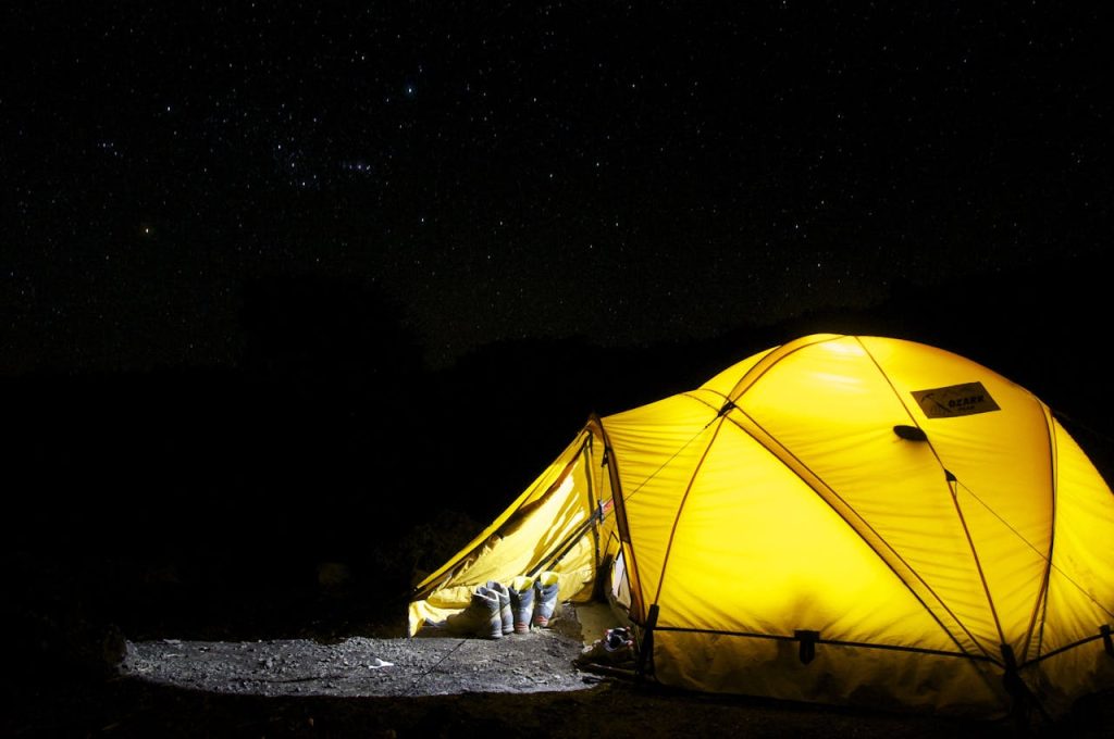 A glowing yellow tent is set up at night under a starry sky, with a few pairs of shoes placed at its entrance. The surrounding area is dark, highlighting the illuminated tent.