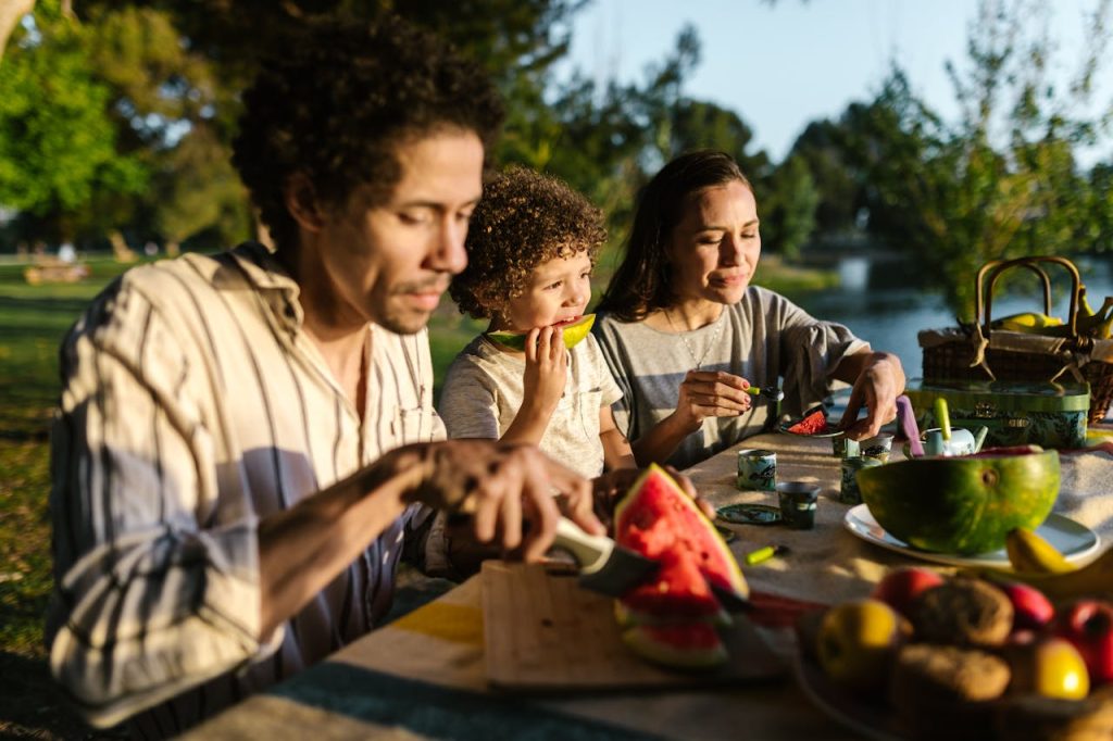 A family of three sits at a picnic table outdoors, enjoying watermelon and other food together on a sunny day, surrounded by trees and a park setting.