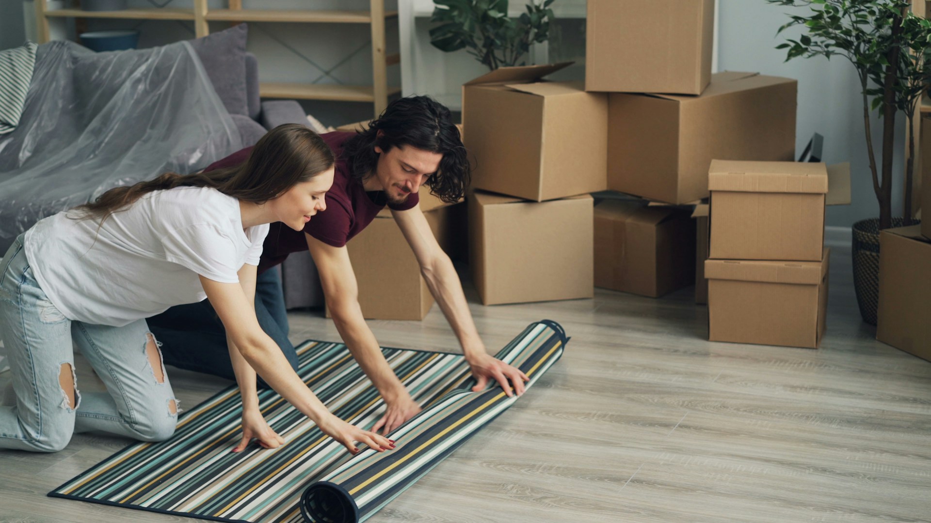 A man and woman kneel on a wooden floor, rolling out a striped rug. Around them are several cardboard moving boxes and some potted plants, suggesting they’re unpacking or moving in.